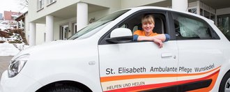 Ein Fahrzeug des Pflegedienst Wunsiedel steht vor einem Gebäude. Aus dem geöffnetem Fahrerfenster lächelt eine Mitarbeiterin des Pflegedienstes Wunsiedel.